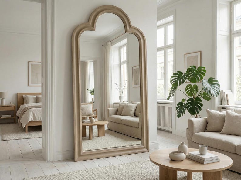 May include: A large, arched-top mirror with a light wood frame stands against a white wall. The mirror reflects a living room with a sofa, coffee table, and a large potted plant. The room has a neutral color palette.