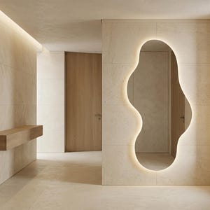 May include: A modern interior featuring a large, organically shaped mirror with a glowing outline. The walls and floor are a light beige, and a wooden bench is mounted on the wall. A wooden door is visible in the background.