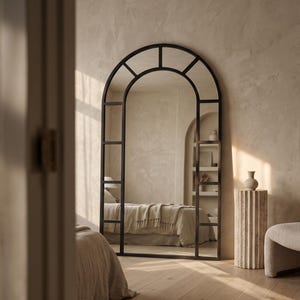May include: A large, floor-standing arched mirror with a dark metal frame divided into rectangular panes. The mirror features a rounded top section and a grid-like structure, reflecting a bedroom interior with a bed and a shelf unit.