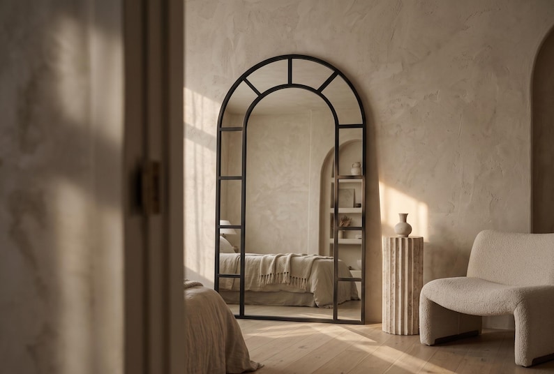 May include: A large, floor-standing arched mirror with a dark metal frame divided into rectangular panes. The mirror features a rounded top section and a grid-like structure, reflecting a bedroom interior with a bed and a shelf unit.