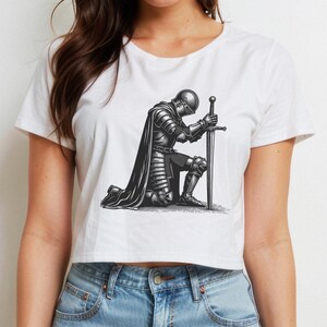 May include: A white cropped t-shirt with a black and white illustration of a knight kneeling with a sword. The knight is wearing armor and a cape.