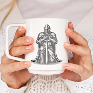 May include: White ceramic mug with a black and white illustration of a knight kneeling with a sword. The knight is wearing armor and a helmet.