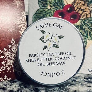 May include: A round metal tin labeled "SALVE GAL" with a floral design. The label lists ingredients: parsley, tea tree oil, shea butter, coconut oil, and beeswax. The tin is 2 ounces and is set against a festive background.