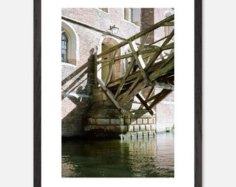 Op de afbeelding: Een houten brug over een kanaal, met een bakstenen gebouw op de achtergrond. De brug is gemaakt van ruw gehouwen balken en heeft een rustieke, verweerde uitstraling. Het kanaal is kalm en reflecterend, en het zonlicht werpt lange schaduwen op het metselwerk.