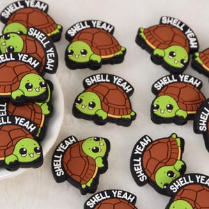 Könnte beinhalten: Eine Sammlung von Cartoon-Schildkröten-Anhängern mit braunen Panzern und grünen Körpern. Jeder Anhänger trägt den Schriftzug "SHELL YEAH" in weißer Schrift auf schwarzem Hintergrund. Die Anhänger liegen auf einer weißen Oberfläche.