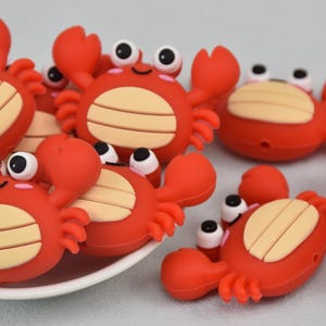 May include: A group of red silicone crab-shaped beads with white eyes and a beige shell. The beads are arranged on a white plate.