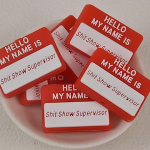 May include: A collection of red and white name tags with the text "HELLO MY NAME IS" and "Shit Show Supervisor" printed on them. The name tags are rectangular and are in a white bowl.