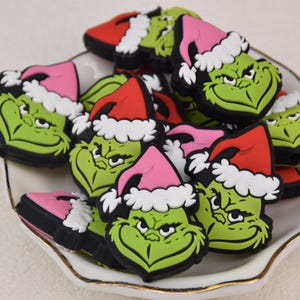 Op de afbeelding: Een verzameling groene Grinch-gezicht bedels, elk met een kerstmuts in rood of roze met witte rand. De bedels zijn gerangschikt in een wit schaaltje met een gouden rand, wat een feestelijk vakantiethema creëert.