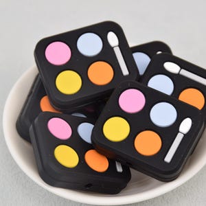 May include: A collection of black, square-shaped makeup palettes with colorful eyeshadows in pink, blue, orange, and yellow. Each palette includes a white applicator. The palettes are arranged in a white bowl.