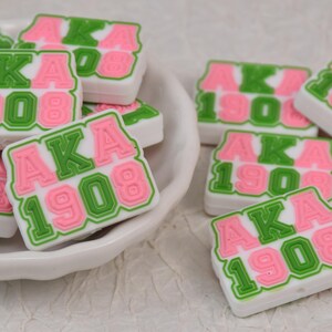 Puede incluir: Cuentas rectangulares blancas con las letras "AKA" en rosa y el número "1908" en verde. Las cuentas están en un cuenco blanco y esparcidas sobre una superficie blanca. Las letras y los números están delineados en verde.