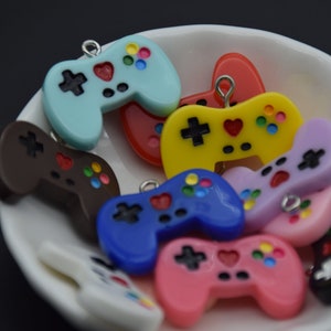 May include: A collection of colourful plastic game controller charms in various colours, including blue, red, yellow, brown, pink, purple, and white. The charms are shaped like video game controllers and have small details like buttons and a joystick.