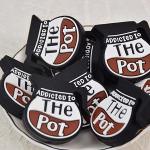 Pode incluir: Uma coleção de pingentes pretos e brancos em forma de cafeteira com o texto "Addicted to The Pot" em um design marrom e branco. Os pingentes estão dispostos em um prato branco.