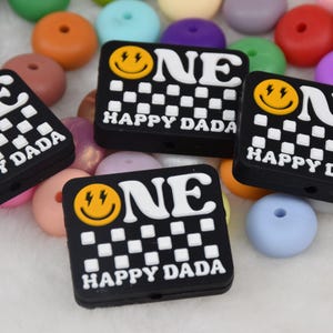 May include: Four black silicone beads with a white checkerboard pattern and a yellow smiley face with lightning bolts. Each bead says "ONE HAPPY DADA" in white letters.