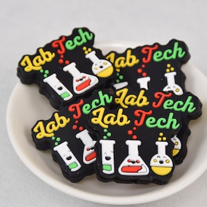 Puede incluir: Un conjunto de cinco dijes de zapatos de goma negra con el texto "Lab Tech" y un gráfico de vasos de precipitados y tubos de ensayo. Los dijes son amarillos, verdes y rojos.