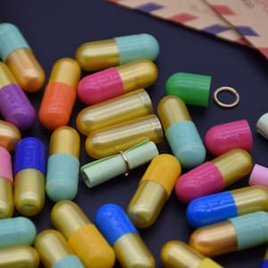 May include: A collection of colorful capsules with gold caps, some containing rolled up paper notes. The capsules are scattered on a black surface.