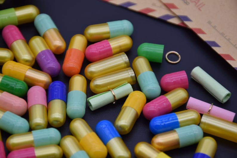 May include: A collection of colorful capsules with gold caps, some containing rolled up paper notes. The capsules are scattered on a black surface.