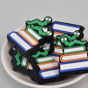 May include: A collection of colorful, cartoonish book-shaped charms with a green caterpillar on top. The books have white, blue, and orange accents. The charms are in a white bowl against a light gray background.