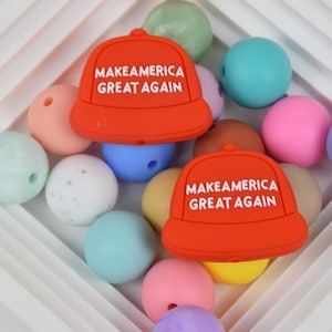 May include: Two red silicone baby teethers shaped like baseball caps with the text "Make America Great Again" in white. The teethers are surrounded by colorful silicone beads.