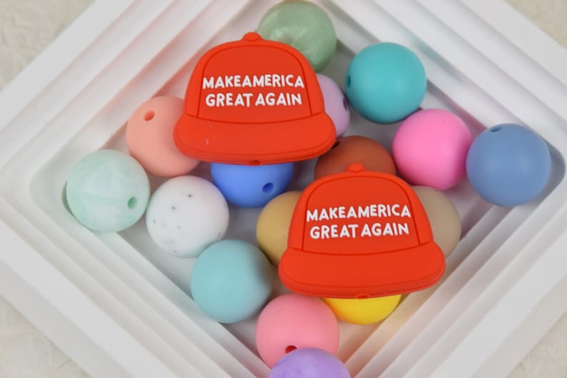 May include: Two red silicone baby teethers shaped like baseball caps with the text "Make America Great Again" in white. The teethers are surrounded by colorful silicone beads.