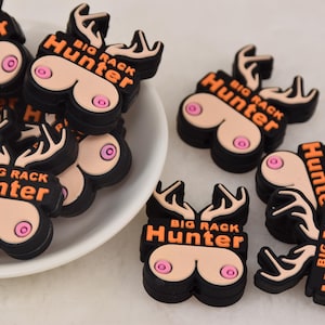 Puede incluir: Una colección de dijes de goma negra y naranja con forma de cabeza de ciervo con cuernos. Cada dije tiene el texto "BIG RACK Hunter".