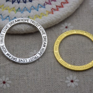 Op de afbeelding: Twee metalen ringen met de woorden "TRUST LOVE DREAM HOPE TRUST" gegraveerd rond de buitenkant. De ring aan de linkerkant is zilver en de ring aan de rechterkant is goud.