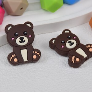 May include: Two brown silicone bear-shaped beads with white and pink details. The beads are designed for teething babies.