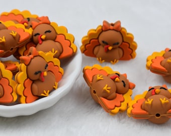 May include: A collection of brown turkey-shaped silicone beads featuring orange and yellow tail feathers, closed eyes, and red wattles. Several beads are clustered inside a white bowl, while others are scattered on a white surface.