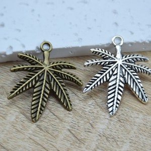 Könnte beinhalten: Zwei Metall-Cannabisblatt-Anhänger, einer in Antikgold und einer in Silber. Beide Anhänger haben ein detailliertes Design und eine Öse zum Anbringen an Schmuck.