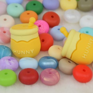 Puede incluir: Una colección de cuentas de silicona de colores con dos cuentas en forma de tarro de miel que dicen "Hunny".