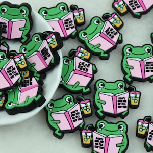 Op de afbeelding: Een verzameling groene kikkerbedels, elk met een roze boek en een drankje. Op het boek staat "DON'T BOTHER ME I'LL EAT YOU". De bedels hebben zwarte contouren en witte ogen. Sommige liggen in een witte kom.