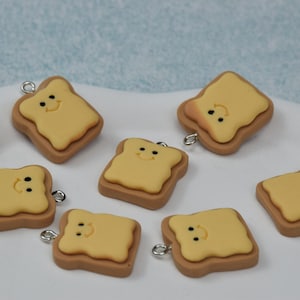 May include: Seven miniature, smiling toast charms with a light brown crust and yellow butter topping. Each charm has a silver-colored loop for attaching to jewelry or other crafts. The charms are arranged on a white surface.