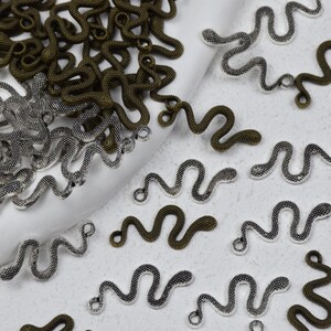 May include: A collection of small, snake-shaped metal charms in antique bronze and silver tones. Each charm features a textured, winding body with a small loop at the head for attachment, scattered across a white surface in various orientations.