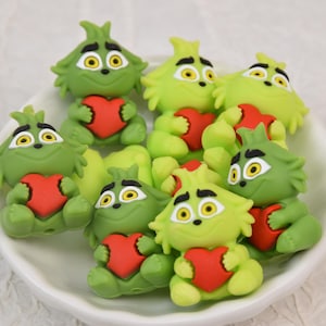 May include: A collection of green figurines resembling a cartoon character, each holding a red heart. The figurines have yellow eyes and are arranged in a white bowl. The overall theme is festive and whimsical.