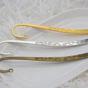 Può includere: Tre segnalibri in metallo dorato, argentato e bronzato, ciascuno con un gancio curvo e un motivo floreale. I segnalibri sono esposti su un piatto bianco oblungo.