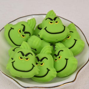 Könnte beinhalten: Eine Sammlung grüner, Grinch-förmiger Verzierungen mit schwarzen und gelben Details, angeordnet in einer weiß-goldfarbenen Schale. Die Grinch-Gesichter haben ein schelmisches Grinsen und eine gerunzelte Stirn.