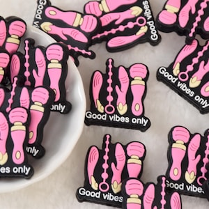 Könnte beinhalten: Eine Sammlung schwarzer und rosa Artikel, einige in einer weißen Schüssel. Die Artikel weisen eine Vielzahl von Formen und Designs auf, wobei auf einigen der Satz "Good vibes only" aufgedruckt ist. Die Artikel sind auf einer hellen Oberfläche verstreut.