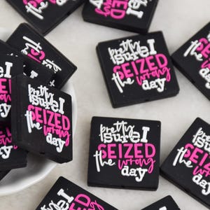 May include: Black square-shaped accessories with the text "Pretty sure I seized the wrong day" in white and pink. Some are in a white bowl, others scattered on a surface.
