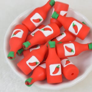 Peut inclure: Un ensemble de petites perles rouges en forme de bouteilles de sauce piquante, avec des bouchons verts et des étiquettes blanches avec un piment rouge. Les perles sont empilées dans un bol blanc.