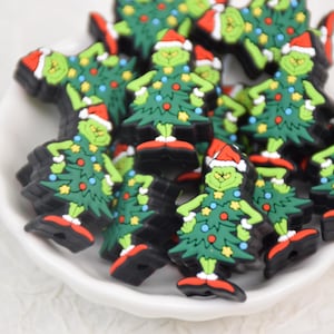 May include: A collection of Grinch-themed Christmas charms. Each charm features the Grinch's face with a Santa hat, a green Christmas tree body, and red shoes. The charms are black around the edges and are in a white bowl.