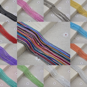 May include: Assortment of 12 colors of ball chain, each chain is made of small metal balls. The chains are laid out in rows on a white fabric background with small white flowers.