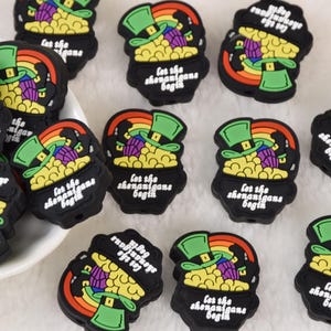 May include: Black rubber charms featuring a leprechaun's hat, a pot of gold, and a rainbow. The charms have the text "Let the shenanigans begin." These charms are perfect for St. Patrick's Day crafts.