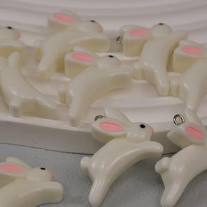 Könnte beinhalten: Elfenbeinfarbene Hasenanhänger mit rosa Innenohren und schwarzen Augen. Diese Resin-Hasenanhänger haben eine silberfarbene Schlaufe zum Befestigen an Schmuck oder anderen Bastelarbeiten. Die Anhänger sind in einer spielerischen, springenden Pose.