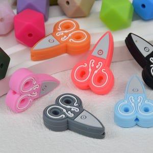 May include: Assortment of colourful silicone baby teethers shaped like scissors. The teethers are in various colours, including pink, orange, blue, grey, and black.
