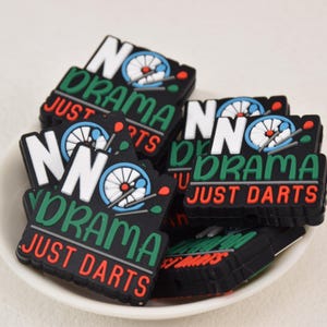 Può includere: Una collezione di toppe rettangolari nere con la scritta "NO DRAMA JUST DARTS" in bianco, verde e rosso. Ogni toppa presenta una grafica del bersaglio delle freccette. Le toppe sono in una ciotola bianca.