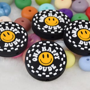 May include: Four black silicone teething beads with a white dashed border and the text "One Happy Dude" in white with a yellow smiley face in the center.