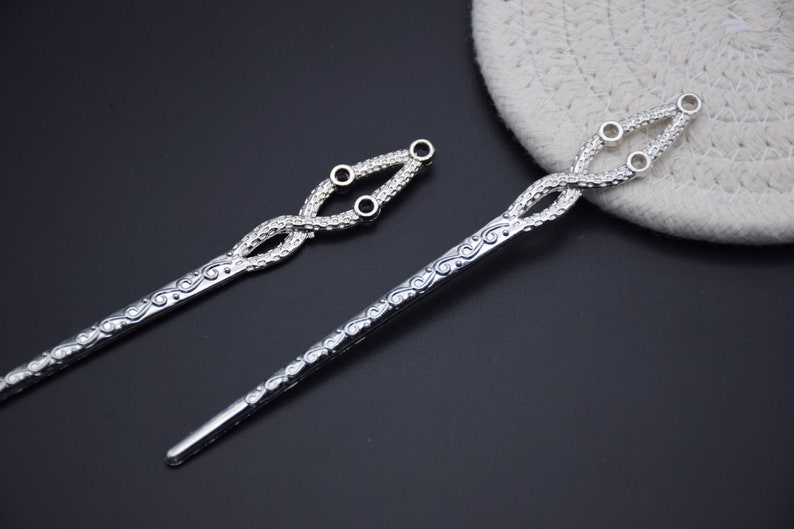 May include: Two silver hair sticks with intricate designs. One stick has a decorative knot at the top with three small holes. The other stick has a more simple design.