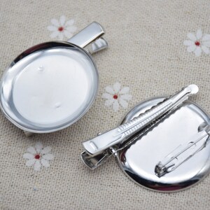 May include: Two silver-toned round metal brooch bases with attached clasps. The bases are blank and ready to be customized.