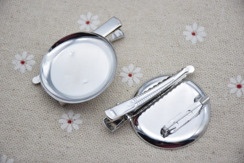 May include: Two silver-toned round metal brooch bases with attached clasps. The bases are blank and ready to be customized.