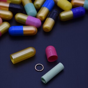 May include: A variety of colorful capsules, some open, on a black surface. The capsules are gold, blue, pink, green, and black.