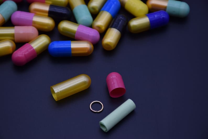 May include: A variety of colorful capsules, some open, on a black surface. The capsules are gold, blue, pink, green, and black.